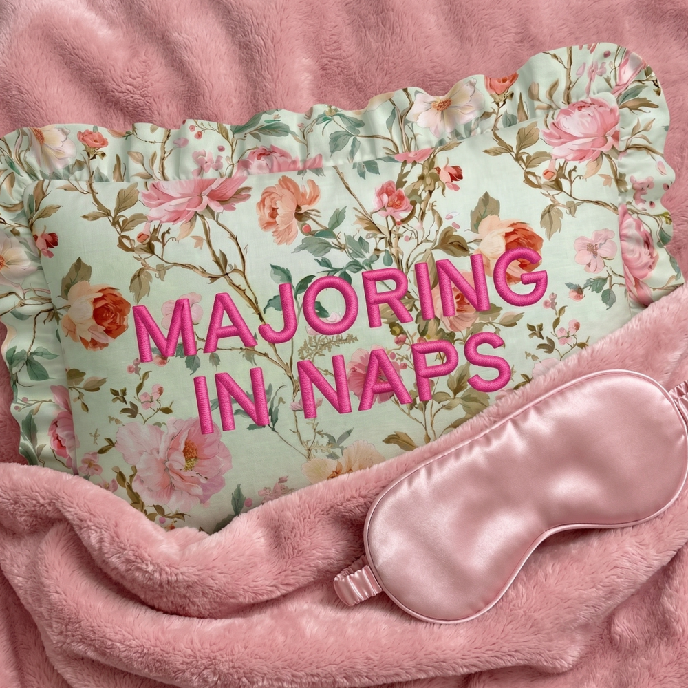 Floral pillow with 'MAJORING IN NAPS' text and pink sleep mask on pink fabric background