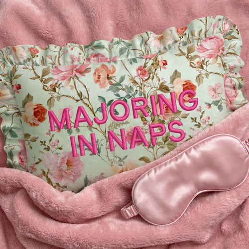Floral pillow with 'MAJORING IN NAPS' text and pink sleep mask on pink fabric background